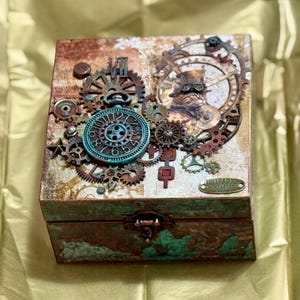 Puede incluir: Una caja cuadrada de madera con acabado envejecido en cobre y verde azulado, decorada con un conjunto de engranajes met&aacute;licos, piezas de reloj y un dial circular en la tapa. Un peque&ntilde;o cierre de tono lat&oacute;n asegura la parte frontal de la caja.