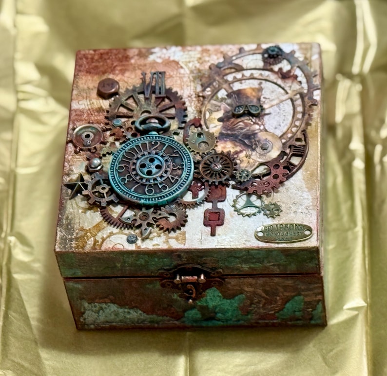 Puede incluir: Una caja cuadrada de madera con acabado envejecido en cobre y verde azulado, decorada con un conjunto de engranajes met&aacute;licos, piezas de reloj y un dial circular en la tapa. Un peque&ntilde;o cierre de tono lat&oacute;n asegura la parte frontal de la caja.