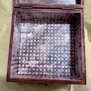 Puede incluir: Una caja cuadrada de madera con un acabado exterior en tono cobre oscuro y desgastado. La base y los lados interiores est&aacute;n forrados con un papel decorativo que presenta un patr&oacute;n de cuadr&iacute;cula en blanco y marr&oacute;n oscuro con escritura cursiva.