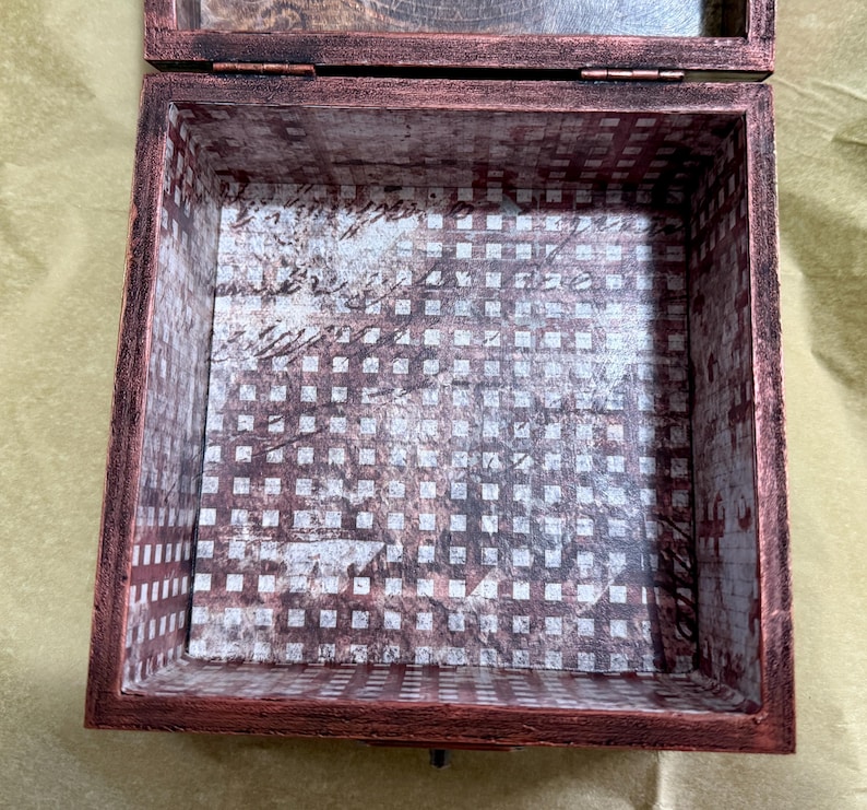 Puede incluir: Una caja cuadrada de madera con un acabado exterior en tono cobre oscuro y desgastado. La base y los lados interiores est&aacute;n forrados con un papel decorativo que presenta un patr&oacute;n de cuadr&iacute;cula en blanco y marr&oacute;n oscuro con escritura cursiva.