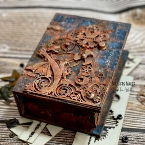 Könnte beinhalten: Eine rechteckige Holzkiste mit Steampunk-Design. Die Kiste ist mit einem verwitterten blau-braunen Finish bemalt und verfügt über kupferfarbene Zahnräder und das Wort "IMAGINE". The Gifts Maker by Ana Webber ist auf der Seite aufgedruckt.
