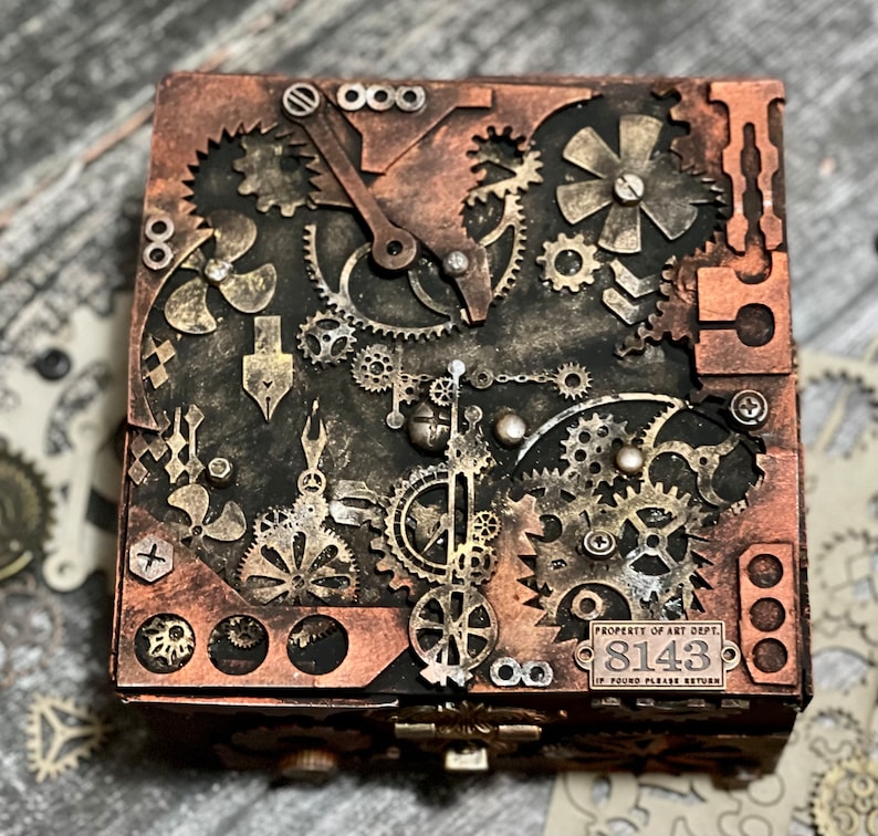 Puede incluir: Una caja cuadrada con un acabado oscuro y desgastado, decorada con un surtido de engranajes, ruedas dentadas y piezas mec&aacute;nicas en tonos cobre y lat&oacute;n. Una peque&ntilde;a placa met&aacute;lica rectangular en el frente muestra el texto PROPERTY OF ART DEPT. 8143 IF FOUND PLEASE RETURN.