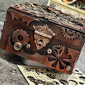 Puede incluir: Una caja de madera de color marr&oacute;n oscuro con un cierre met&aacute;lico decorativo y varios adornos de engranajes y ruedas dentadas de color cobre fijados en la parte frontal y la tapa. La caja tiene forma rectangular con tapa abatible.