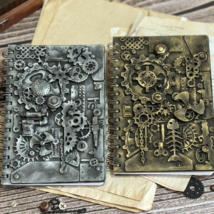 May include: Two spiral-bound notebooks featuring raised, textured covers decorated with an assortment of metallic gears, cogs, mechanical parts, and a fish skeleton. One notebook has a silver finish, while the other has a bronze finish.