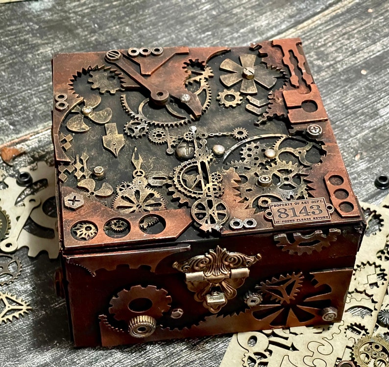 Puede incluir: Una caja cuadrada con acabado oscuro, decorada con numerosos engranajes, ruedas dentadas y componentes mec&aacute;nicos de color cobre. Un cierre de tono lat&oacute;n asegura la parte frontal. Una peque&ntilde;a placa met&aacute;lica arriba a la derecha muestra el texto Property of Art Dept. 8143.