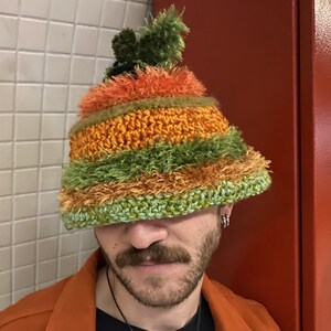 Puede incluir: Un sombrero de ganchillo hecho a mano y colorido. El sombrero presenta rayas horizontales de color naranja, verde y amarillo. La parte superior del sombrero tiene un pompón verde. El sombrero lo lleva una persona.