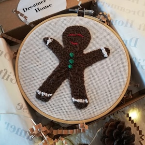 May include: A brown gingerbread man with white buttons and green buttons is embroidered on a white fabric in a wooden embroidery hoop. The hoop is sitting on a brown paper surface with white paper and pine cones.