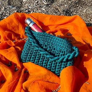 May include: A teal crocheted handbag with a rope-like strap rests on a vibrant orange fabric. A silver and red lipstick tube is partially visible inside the bag. The background features a sandy beach with small pebbles.