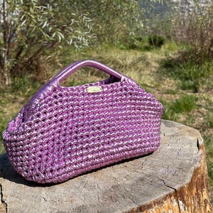 May include: A metallic purple handbag with a woven texture and a matching handle. The bag is sitting on a weathered wooden surface, with a natural outdoor background. A small rectangular tag is attached to the bag.