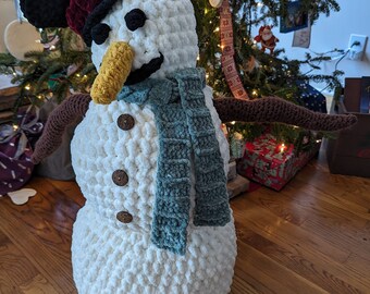 May include: A handmade, plush snowman with a black top hat, burgundy brim, and a yellow carrot nose. It has a teal scarf, brown twig arms, and wooden button details. The snowman is a festive holiday decoration.