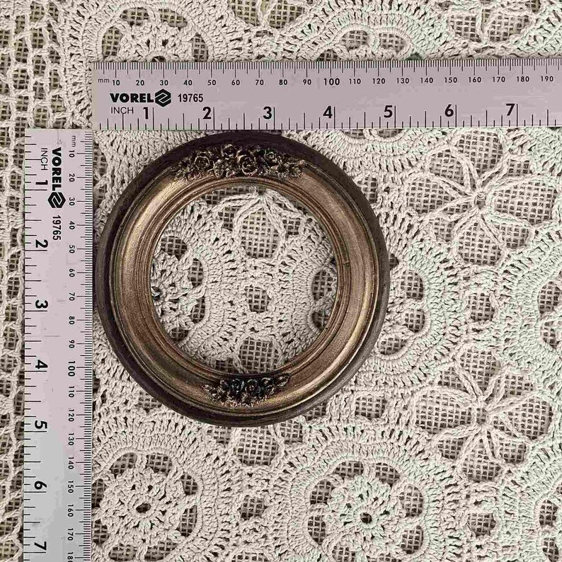 K&ouml;nnte beinhalten: Ein kleiner, runder Bilderrahmen im antiken Stil mit dunkelbraunem Finish und floralen Akzenten. Der Rahmen hat einen Durchmesser von etwa 7.6 cm und liegt auf einer geh&auml;kelten Tischdecke. Ein Lineal liegt neben dem Rahmen.