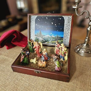 Könnte beinhalten: Eine Holzbox mit Scharnierdeckel, die eine Miniatur-Weihnachtskrippe beherbergt. Die Szene umfasst Figuren von Maria, Josef, dem Jesuskind, Engeln und den drei Weisen, vor einer gemalten Kulisse einer sternenklaren Nacht und einer fernen Stadt.