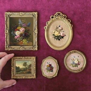 Op de afbeelding: Een verzameling kleine, sierlijke miniatuurschilderijen met gouden lijst. De schilderijen tonen bloemstillevens en landschappen. De lijsten hebben ingewikkelde details en zijn geplaatst op een dieprode achtergrond.