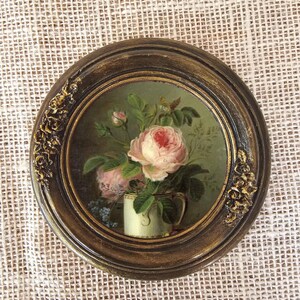 K&ouml;nnte beinhalten: Gerahmtes rundes Stillleben mit Blumen. Das Gem&auml;lde zeigt einen Strau&szlig; rosa Rosen in einer wei&szlig;en Vase vor gr&uuml;nem Hintergrund. Der Rahmen ist dunkelbraun mit goldenen Akzenten.