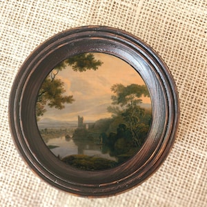 Può includere: Dipinto di paesaggio ovale incorniciato, raffigurante un castello in riva a un lago. Il dipinto è racchiuso in una cornice di legno marrone scuro. L'opera raffigura alberi, acqua e un cielo nuvoloso.