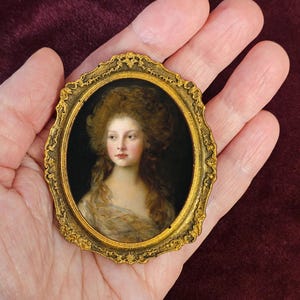 Può includere: Un ritratto in miniatura ovale di una donna con capelli castani ricci, in una cornice dorata ornata. Il ritratto mostra una donna con un incarnato pallido, che indossa un abito di colore chiaro. La cornice è tenuta in mano su uno sfondo rosso scuro.