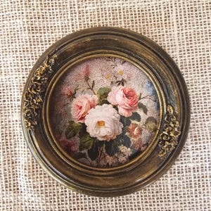 K&ouml;nnte beinhalten: Ein runder, gerahmter Blumendruck im Vintage-Stil. Der Druck zeigt einen Strau&szlig; rosa und wei&szlig;er Rosen mit gr&uuml;nen Bl&auml;ttern vor einem weichen, strukturierten Hintergrund. Der Rahmen ist dunkelgoldfarben mit aufwendigen Details.