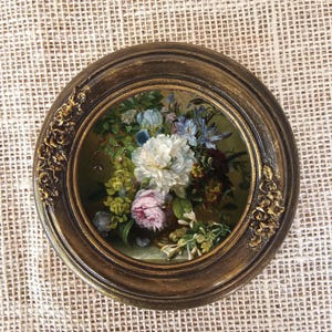 K&ouml;nnte beinhalten: Gerahmtes rundes Blumenbild mit einem Strau&szlig; aus wei&szlig;en, rosa und blauen Blumen. Der dunkelbraune Rahmen hat goldene Akzente und florale Verzierungen. Das Kunstwerk wird auf einer strukturierten, cremefarbenen Oberfl&auml;che pr&auml;sentiert. Der Durchmesser des Rahmens betr&auml;gt ca. 20 cm.
