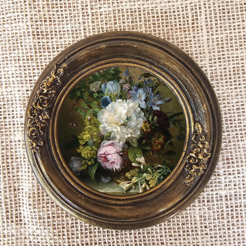 K&ouml;nnte beinhalten: Gerahmtes rundes Blumenbild mit einem Strau&szlig; aus wei&szlig;en, rosa und blauen Blumen. Der dunkelbraune Rahmen hat goldene Akzente und florale Verzierungen. Das Kunstwerk wird auf einer strukturierten, cremefarbenen Oberfl&auml;che pr&auml;sentiert. Der Durchmesser des Rahmens betr&auml;gt ca. 20 cm.