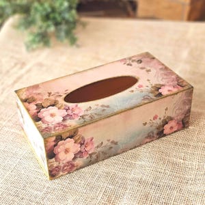 May include: A rectangular tissue box cover featuring a soft pink and blue background with a floral pattern of pink roses and green leaves. The box has a central oval opening on the top and gold-toned edges along the corners and base.