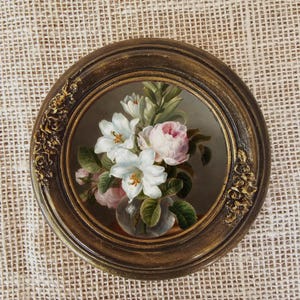 K&ouml;nnte beinhalten: Gerahmtes, rundes Blumenbild mit wei&szlig;en und rosa Bl&uuml;ten in einer Glasvase. Der dunkelbraune Rahmen hat goldfarbene Verzierungen. Der Hintergrund ist ein strukturiertes, neutralfarbenes Gewebe.