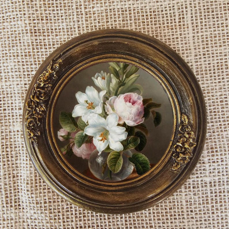K&ouml;nnte beinhalten: Gerahmtes, rundes Blumenbild mit wei&szlig;en und rosa Bl&uuml;ten in einer Glasvase. Der dunkelbraune Rahmen hat goldfarbene Verzierungen. Der Hintergrund ist ein strukturiertes, neutralfarbenes Gewebe.