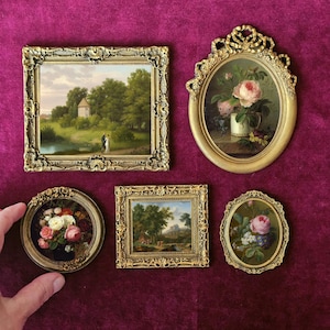 May include: Five miniature framed paintings are displayed. The artworks depict landscapes and floral still lifes, each in an ornate gold-coloured frame. The paintings are presented on a deep red velvet surface.