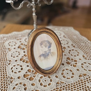 Peut inclure: Un cadre photo ovale doré avec une photo en noir et blanc d'une femme portant un col en dentelle et une robe sombre. Le cadre est posé sur une dentelle blanche crochetée.