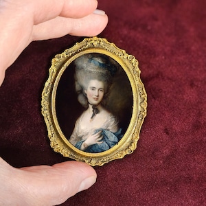 Può includere: Un ritratto in miniatura antico in una cornice ovale dorata e ornata. Il ritratto raffigura una donna con una parrucca impolverata, che indossa un abito bianco e uno scialle blu. La cornice ha un design dettagliato e strutturato.