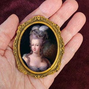 Może przedstawiać: Owalny miniaturowy portret kobiety w złotej ramie. Portret przedstawia kobietę z pudrowanymi włosami, niebieską suknią i wysadzaną klejnotami broszką. Złota rama ma bogaty, szczegółowy wzór.