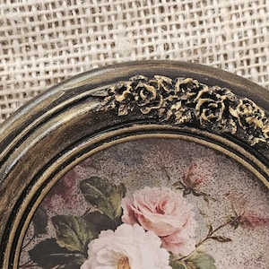 K&ouml;nnte beinhalten: Nahaufnahme eines ovalen Bilderrahmens im Vintage-Stil. Der Rahmen hat eine dunkle, gealterte Goldoberfl&auml;che mit aufwendigen floralen Details an der Oberseite. Das Bild im Inneren zeigt eine weiche Malerei im Aquarellstil mit rosa und wei&szlig;en Rosen.