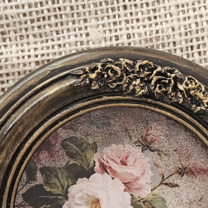 K&ouml;nnte beinhalten: Nahaufnahme eines ovalen Bilderrahmens im Vintage-Stil. Der Rahmen hat eine dunkle, gealterte Goldoberfl&auml;che mit aufwendigen floralen Details an der Oberseite. Das Bild im Inneren zeigt eine weiche Malerei im Aquarellstil mit rosa und wei&szlig;en Rosen.