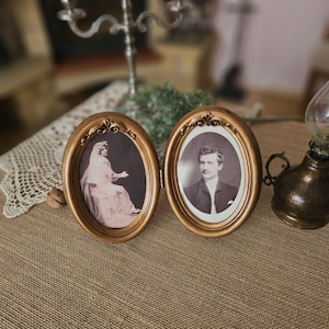 Può includere: Due cornici ovali dorate con dettagli decorativi. Le cornici sono unite e ciascuna contiene una fotografia in bianco e nero di una persona. La cornice a sinistra mostra una donna con un abito bianco e la cornice a destra mostra un uomo in abito e cravatta.
