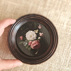 Puede incluir: Un pequeño marco redondo de madera oscura con una naturaleza muerta floral pintada. La pintura presenta un ramo de flores blancas, rosas y rojas sobre un fondo oscuro. El marco está sostenido por una mano.