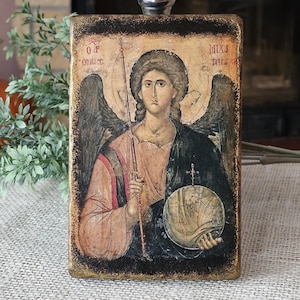 Op de afbeelding: Een handgeschilderd icoon van een engel met vleugels, die een bol en een speer vasthoudt. Het icoon is geschilderd op een houten paneel met een donkerbruine achtergrond en gouden accenten. De engel is afgebeeld met een halo en een lange, vloeiende mantel. De tekst op het icoon luidt "ΟΡ ΟΘΕΟΣ" en "MIXI ΠΗΛΗ".