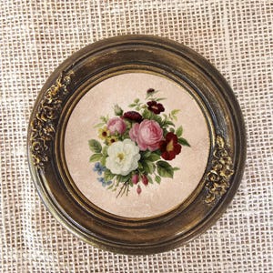 K&ouml;nnte beinhalten: Runder, gerahmter Blumendruck mit einem Bouquet aus rosa, wei&szlig;en und roten Rosen, gr&uuml;nen Bl&auml;ttern und kleinen blauen Bl&uuml;ten. Der dunkelbraune Rahmen hat goldfarbene Akzente und dekorative florale Details. Der Druck ist vor einem hellrosa Hintergrund.