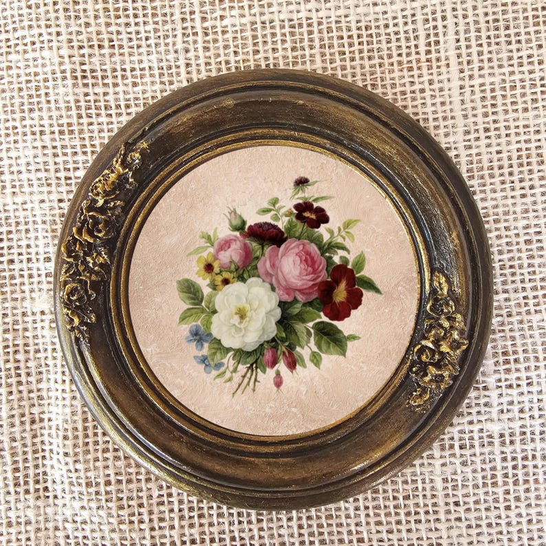 K&ouml;nnte beinhalten: Runder, gerahmter Blumendruck mit einem Bouquet aus rosa, wei&szlig;en und roten Rosen, gr&uuml;nen Bl&auml;ttern und kleinen blauen Bl&uuml;ten. Der dunkelbraune Rahmen hat goldfarbene Akzente und dekorative florale Details. Der Druck ist vor einem hellrosa Hintergrund.