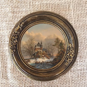 Könnte beinhalten: Rund gerahmtes Gemälde, das eine Winterszene mit einem Schloss, Bäumen und Eisläufern zeigt. Der dunkelbraune Rahmen hat goldfarbene Akzente und florale Verzierungen. Das Gemälde hat eine Vintage-Ästhetik.