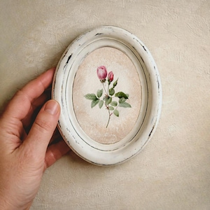 Può includere: Cornice ovale bianca anticata con un acquerello di una rosa rosa e boccioli. L'opera presenta foglie verdi e uno sfondo beige. La cornice è tenuta da una mano.