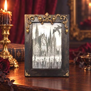 May include: A rectangular black picture frame with ornate gold-colored filigree and skull accents at the top corners and center. The frame holds a black and white illustration of a gothic cathedral, a graveyard with tombstones, and a flying dragon.