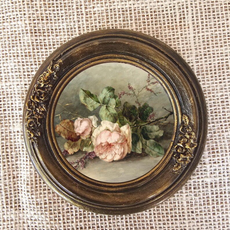K&ouml;nnte beinhalten: Gerahmtes rundes Blumenbild mit rosa und wei&szlig;en Rosen, gr&uuml;nen Bl&auml;ttern und kleinen lila Bl&uuml;ten. Der dunkelbraune Rahmen hat goldfarbene Akzente und dekorative Eckdetails.