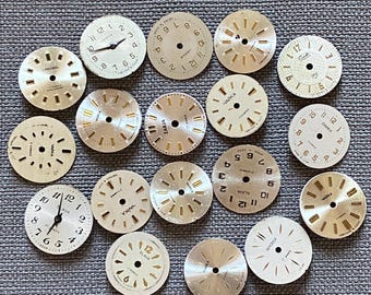 May include: A collection of seventeen circular watch dials in various metallic shades of silver and gold. The dials feature diverse designs, including numerical markers, indices, and brand names such as Slava, Chaika, Zarja, and Sekonda.
