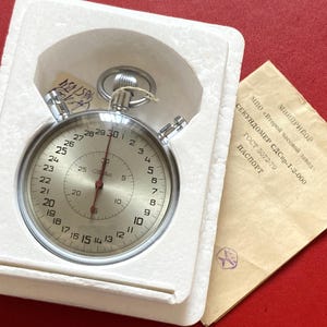 May include: A vintage stopwatch with a silver-colored case and a white face, displayed in a white foam holder. The face has black numerals and a red second hand. A paper document with Cyrillic text is visible.
