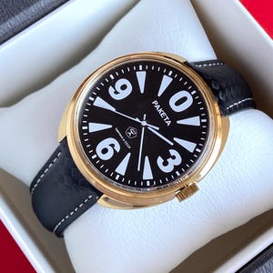 Puede incluir: Un reloj de pulsera dorado con una esfera negra y números blancos. El reloj tiene una correa de cuero negra y dice "PAKEta" en la esfera.