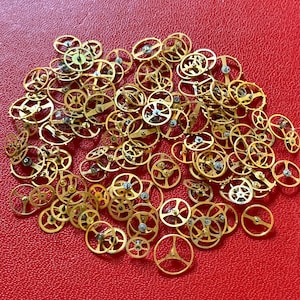 Puede incluir: Una colección de pequeños engranajes de reloj dorados esparcidos sobre una superficie roja. Los engranajes varían en tamaño y diseño, con detalles intrincados visibles. La imagen muestra la precisión y la artesanía de los componentes de relojería.