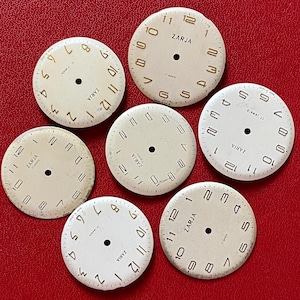 May include: A collection of seven circular off-white watch dials, each featuring the brand name ZARJA, the text 17 jewels, and a central hole. The dials display various numerical or stick marker layouts in black or gold, with some showing signs of age.
