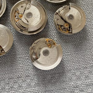 May include: Several disassembled watch movements, revealing detailed mechanical parts. The silver-coloured metal components include gears, springs, and balance wheels, with small gold accents. The watch parts are arranged on a textured grey surface.