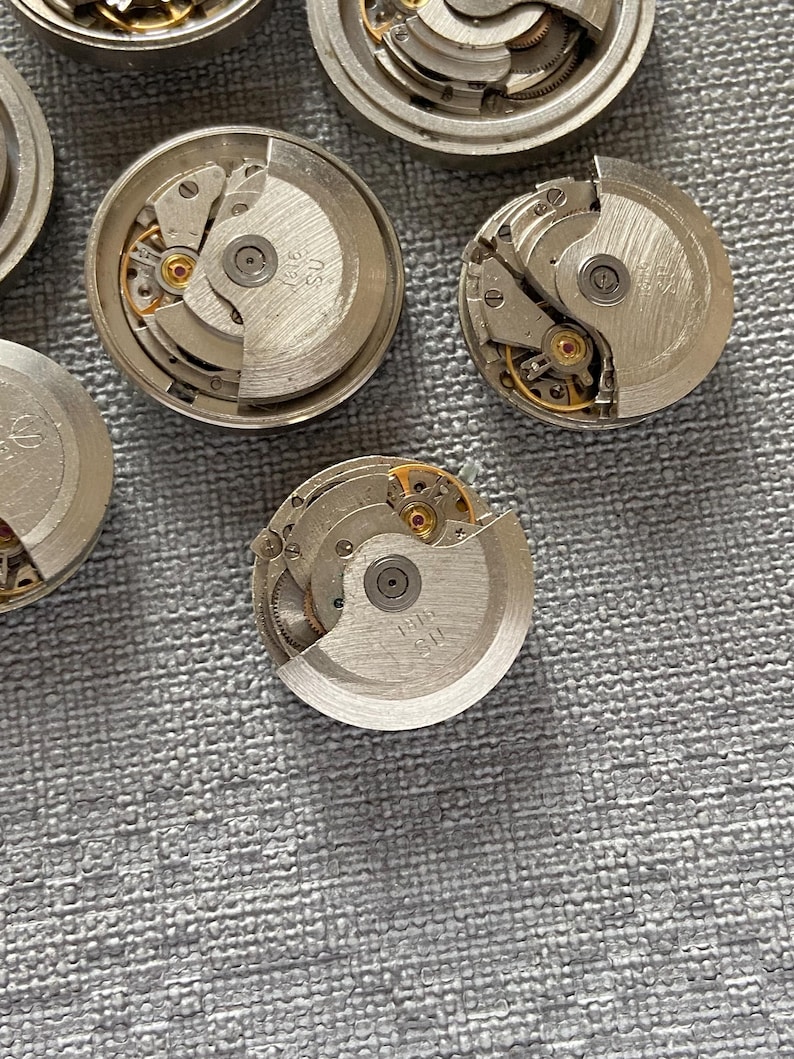May include: Several disassembled watch movements, revealing detailed mechanical parts. The silver-coloured metal components include gears, springs, and balance wheels, with small gold accents. The watch parts are arranged on a textured grey surface.
