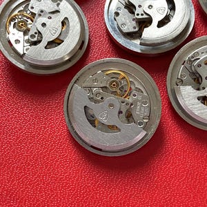 Puede incluir: Primer plano de varios movimientos de reloj desmontados. Los mecanismos plateados presentan intrincados engranajes, joyas y otros componentes. Los movimientos del reloj est&aacute;n dispuestos sobre una superficie roja, mostrando el funcionamiento interno de los relojes.