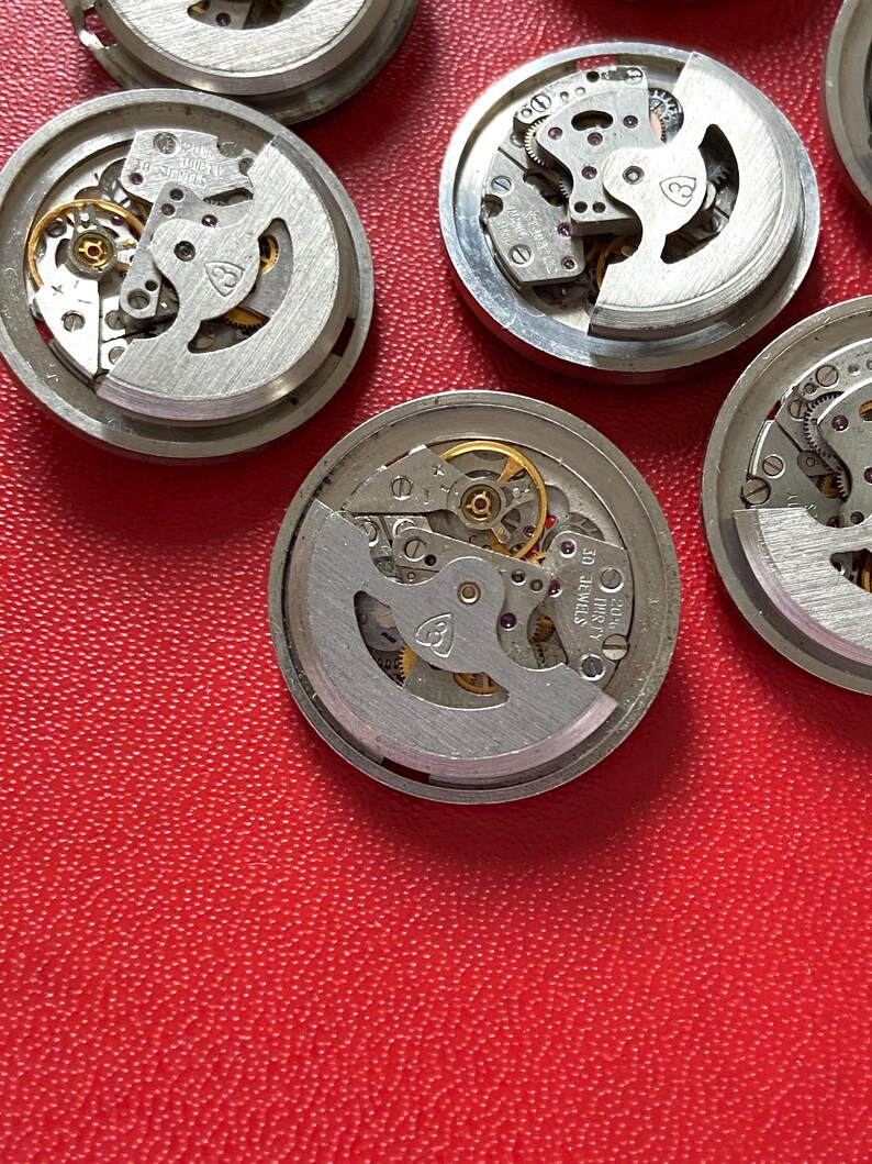 Puede incluir: Primer plano de varios movimientos de reloj desmontados. Los mecanismos plateados presentan intrincados engranajes, joyas y otros componentes. Los movimientos del reloj est&aacute;n dispuestos sobre una superficie roja, mostrando el funcionamiento interno de los relojes.